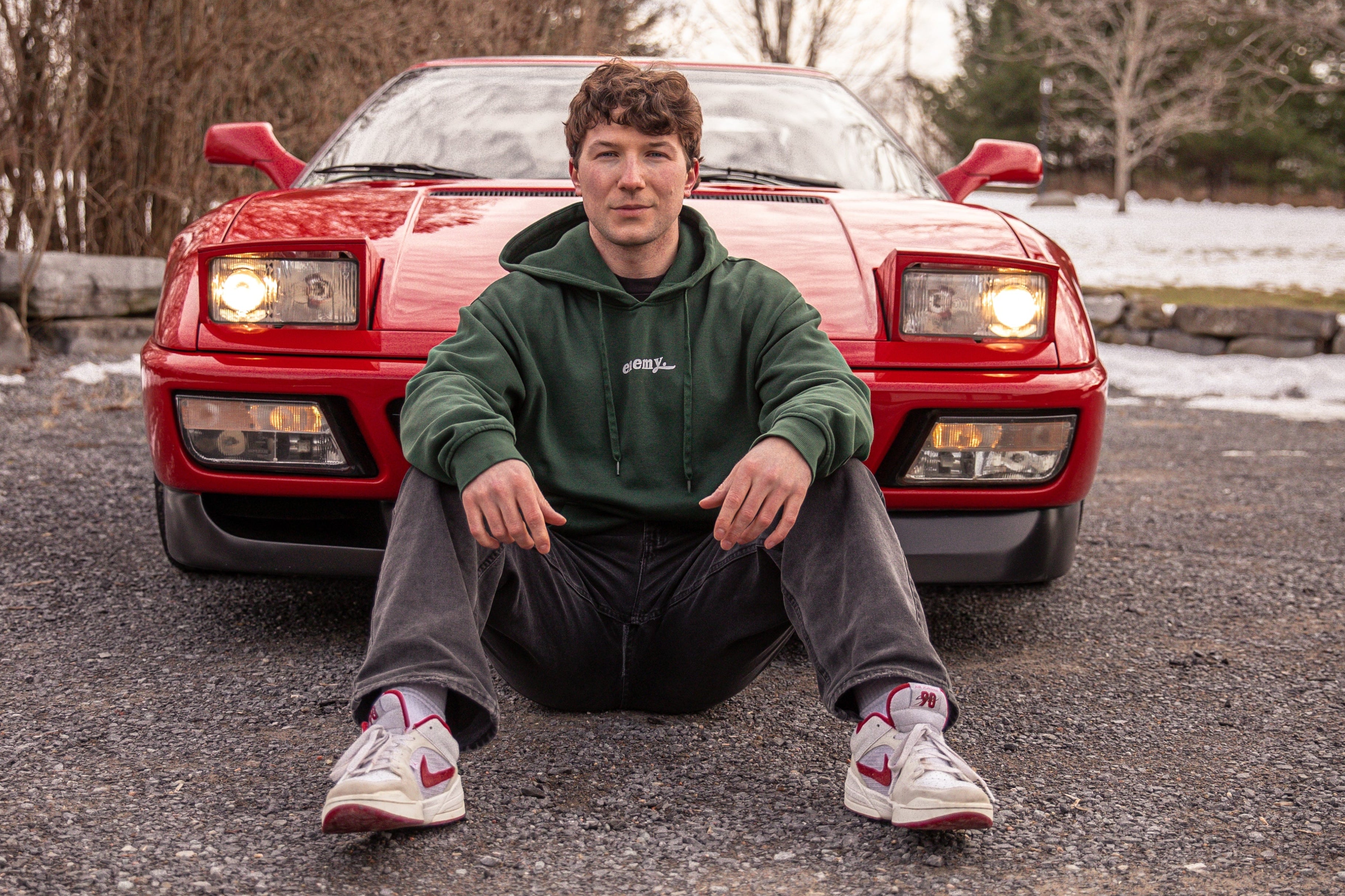 A young man wearing a green hoodie and gray sweatpants sits on the gravel in front of a red sports car, with bare trees and a cloudy sky in the background.